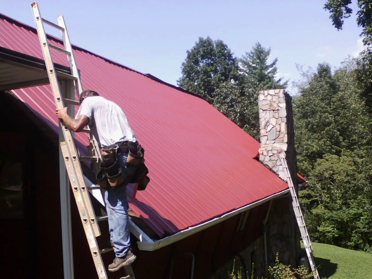 Roofer on a ladder beside a red metal roof and stone chimney — Soffit & Fascia Repair by Roofing Brothers in Gambrills